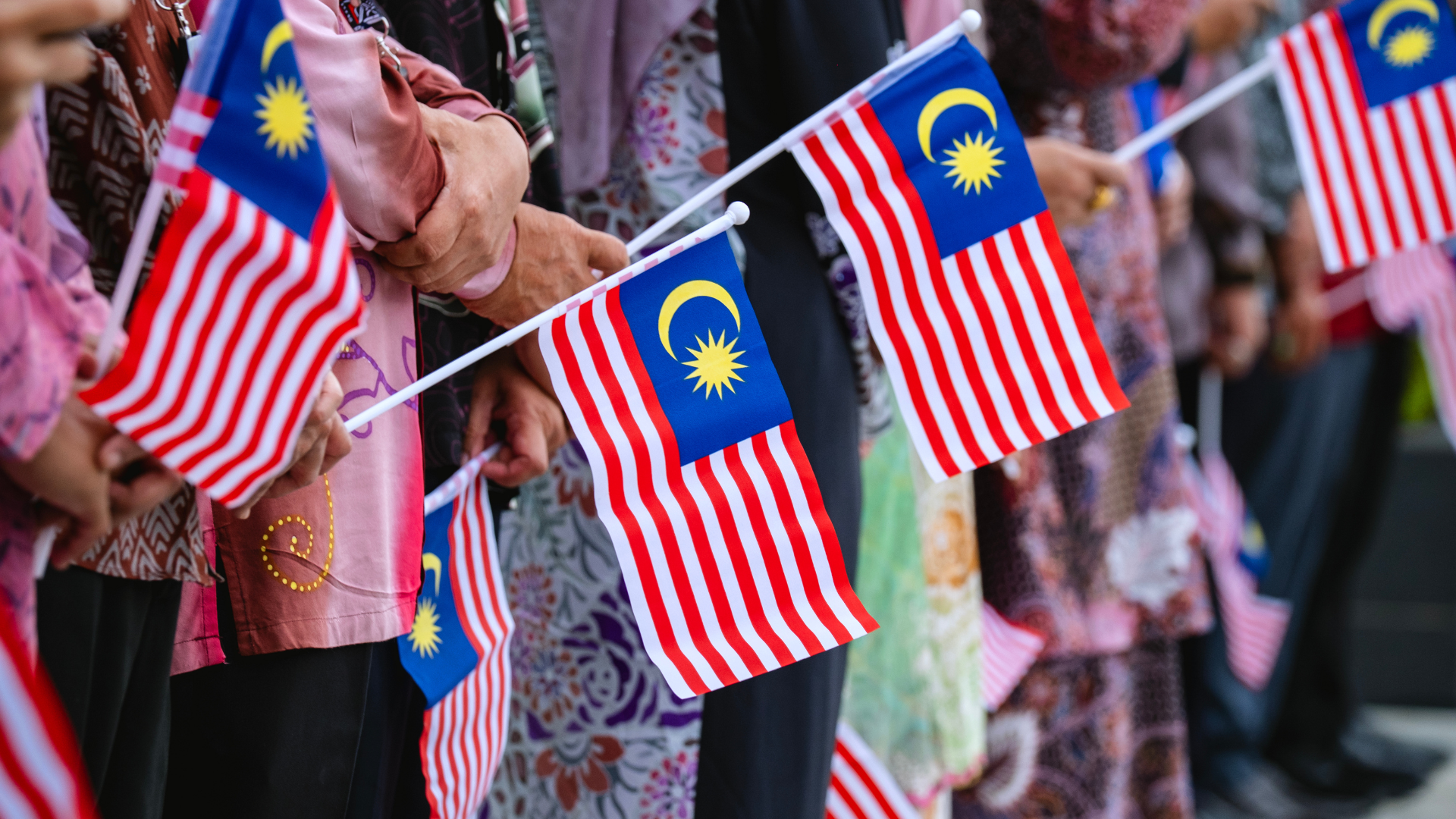 Attendees dressed in diverse traditional and cultural attire holding Jalur Gemilang flags