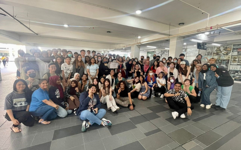 "Large diverse group of smiling Taylor's students posing for a group photo in a spacious outdoor area with a covered walkway