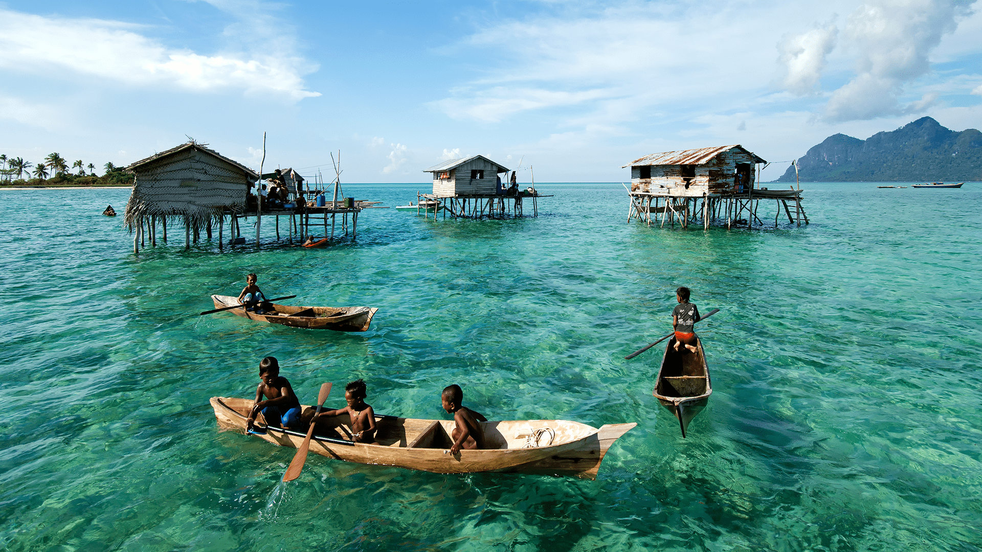 Children paddle wooden canoes amidst stilt houses, while the backdrop reveals lush mountains and clear azure waters, illustrating a harmonious blend of life and nature.