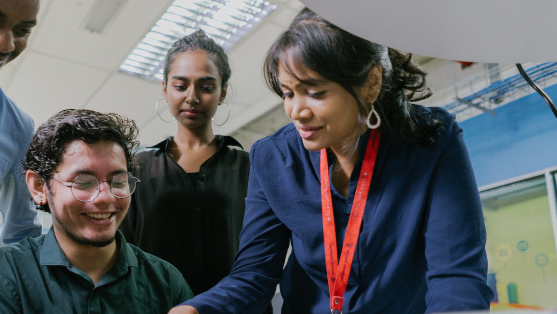A female instructor guides a group of students with a hands-on electronics project in a classroom, all focused on the components on the table.