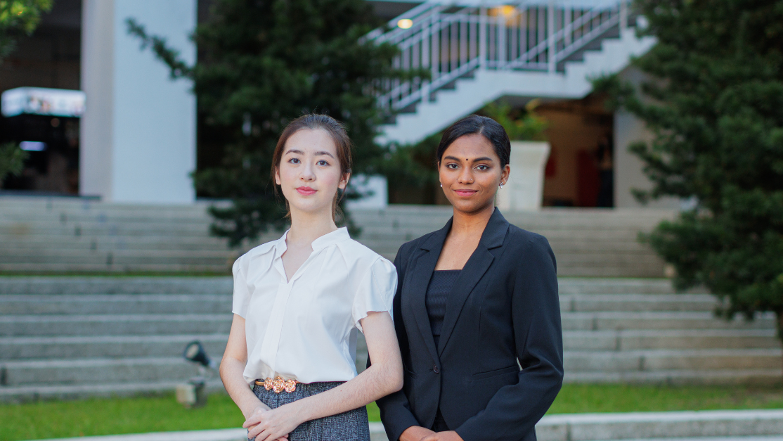 Two professional young women, one in a business suit and the other in smart casual attire, stand confidently side by side on a modern campus.