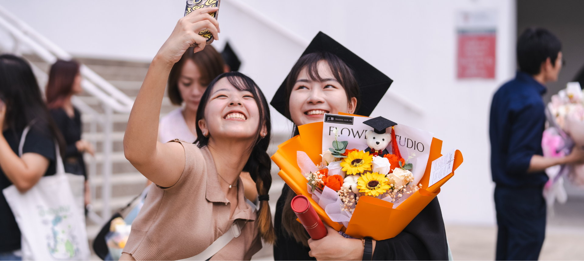 A young woman takes a happy selfie with her friend, who is dressed in a graduation cap and gown and holding a bouquet of flowers.