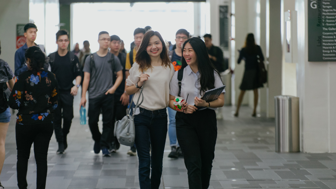 Two female students smile and chat while walking through a busy corridor on campus, with many other students around them.