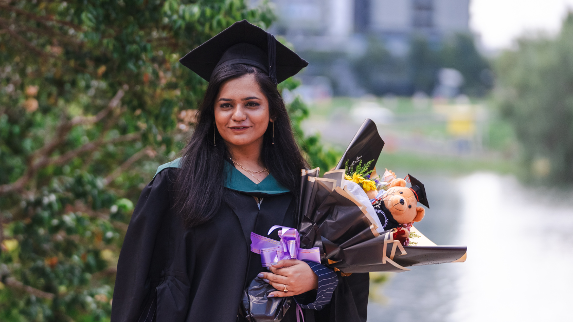 A female graduate in her cap and gown smiles for the camera while holding a bouquet of flowers, with a lake visible behind her.