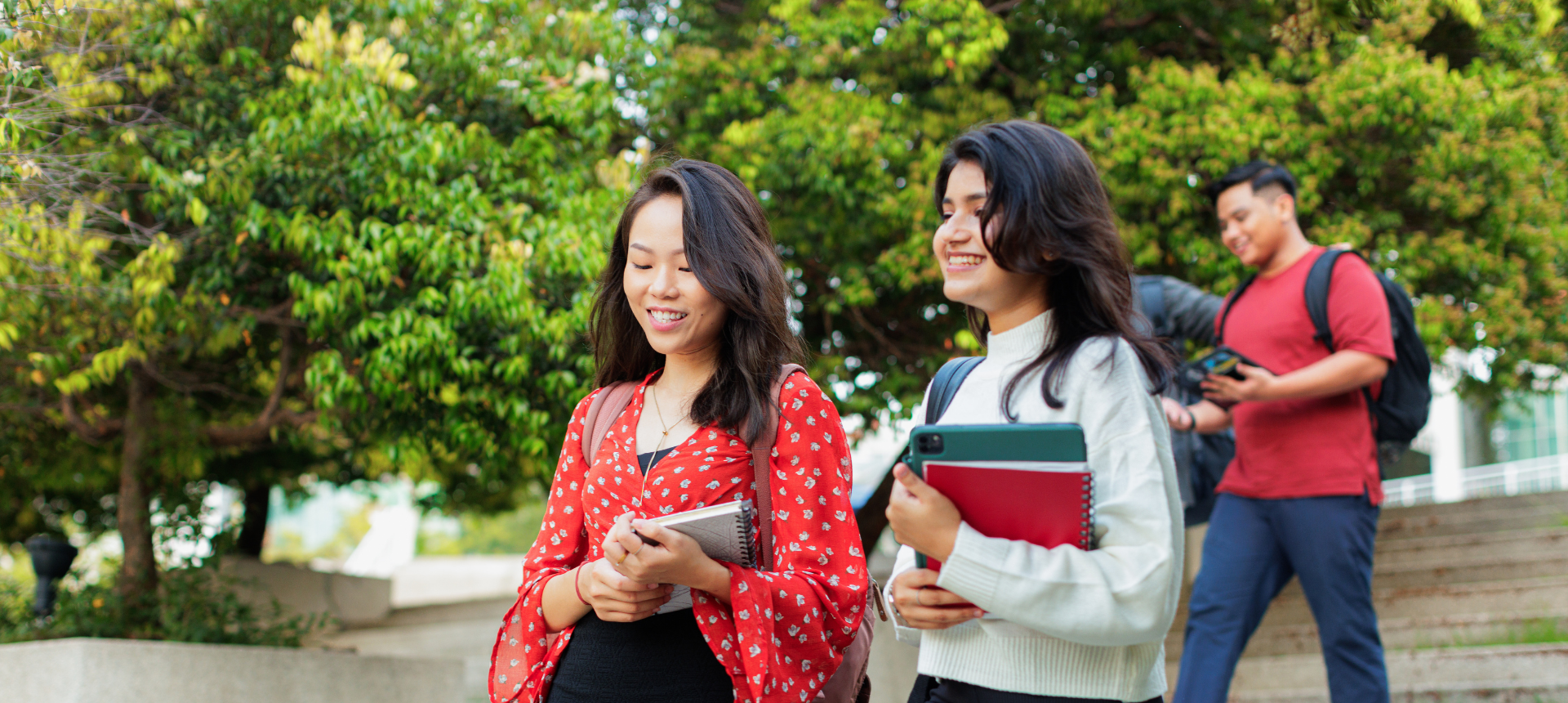Two female university students smile as they walk down stone steps on a leafy campus, carrying their books and notebooks.
