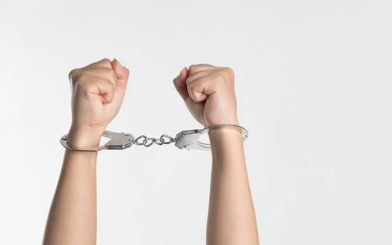 wo hands raised and cuffed together with a pair of metallic handcuffs, set against a white background.