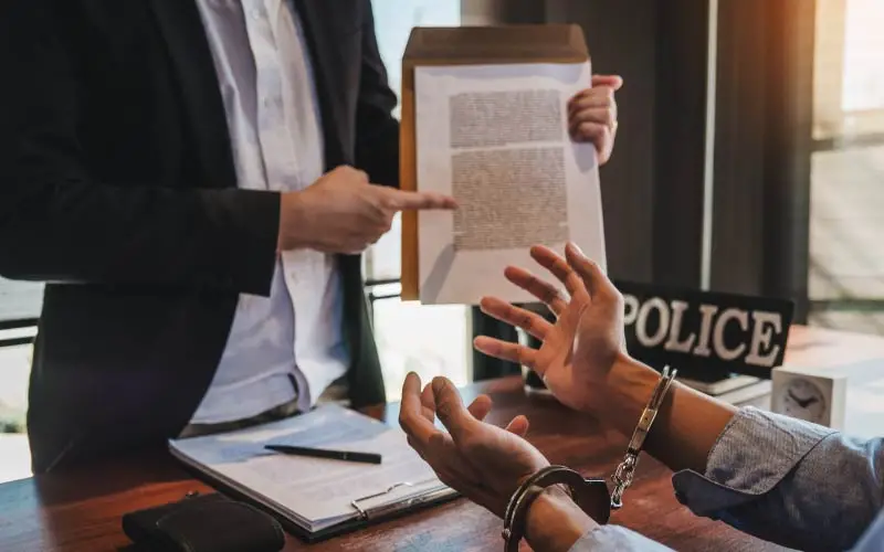A person in handcuffs is engaged in a conversation with another individual pointing to a document, suggesting a legal or official discussion.