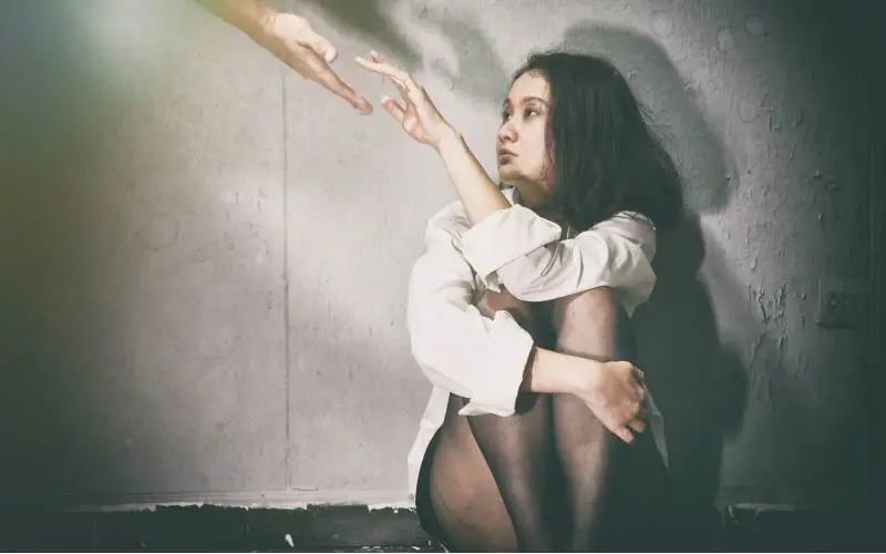 A despondent young person sitting against a wall with knees drawn up, reaching out towards a diffused light source. 