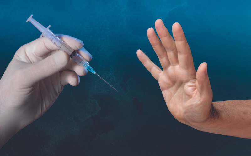 A hand in a medical glove holding a syringe is positioned against a dark blue background, facing a person's hand rejecting the injection, symbolising vaccine hesitancy or refusal.