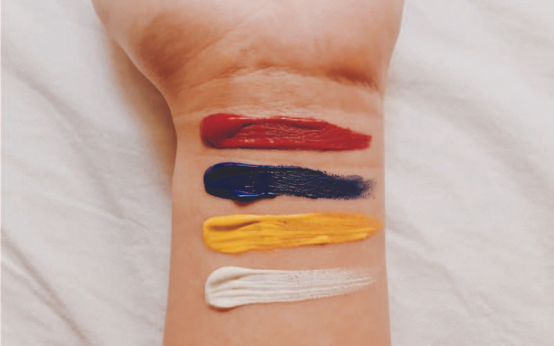 A close-up of a person's forearm with four strokes of paint in red, blue, yellow, and white, symbolising artistic expression of the Malaysian flag colors.