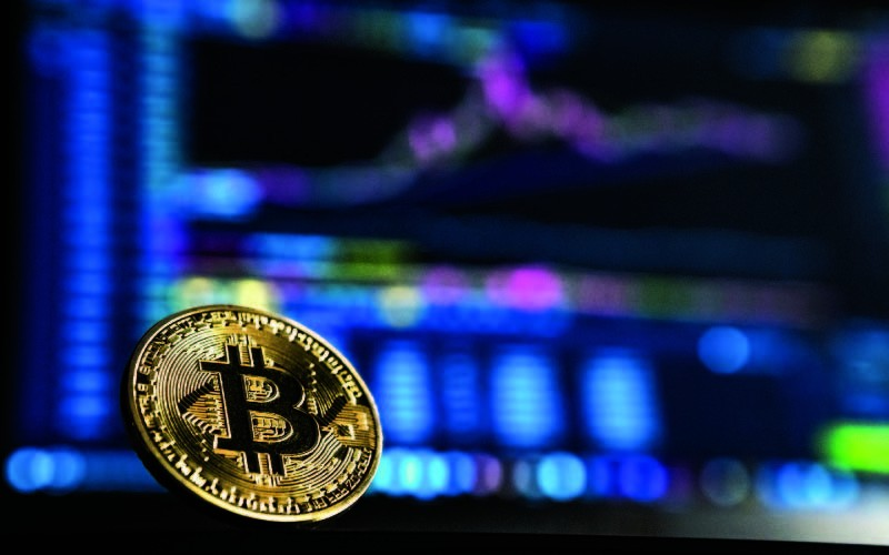 A physical Bitcoin in front of a digital screen displaying financial charts