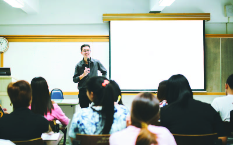 A male lecturer giving a lecture to a class full of Master's students.