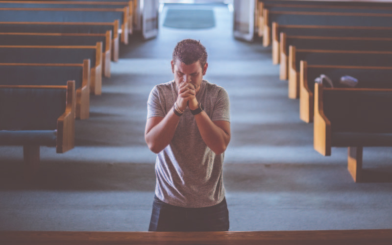 A man, on his knees with his hands clasped together, praying amongst pews.