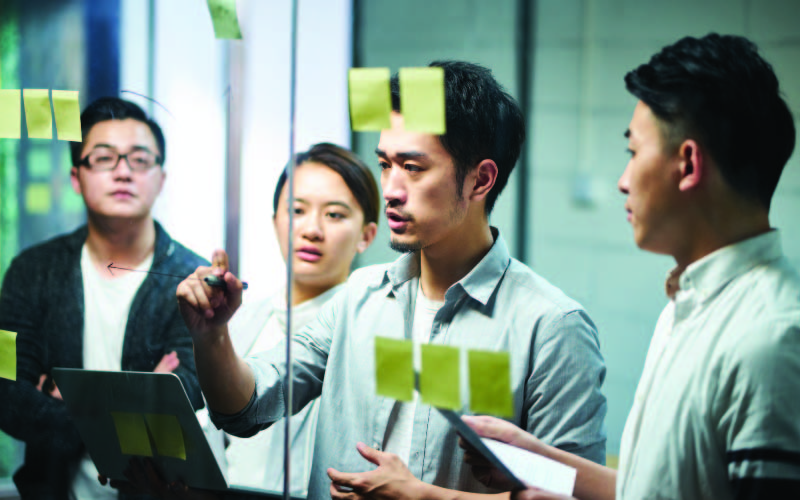 A team of business students brainstorming and writing ideas on a glass wall with sticky notes for a presentation.