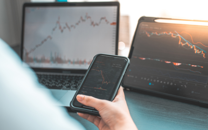 Image depict a hand holding a smartphone, with screen showing the graph of a stock