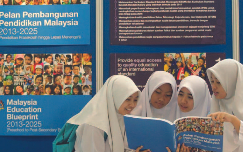 Four young students in hijabs are engrossed in reading a booklet together. They stand in front of informational posters about the 'Malaysia Education Blueprint 2013-2025', which promotes quality education standards.