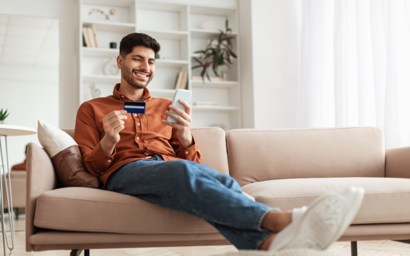 Man sitting on a couch, holding a credit card and a phone in each hand