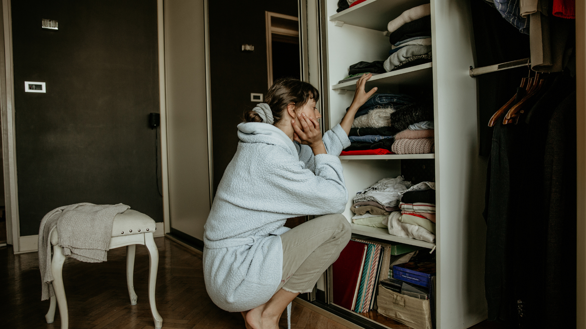 A woman squatting, her hand on her jaw, staring at her open closet