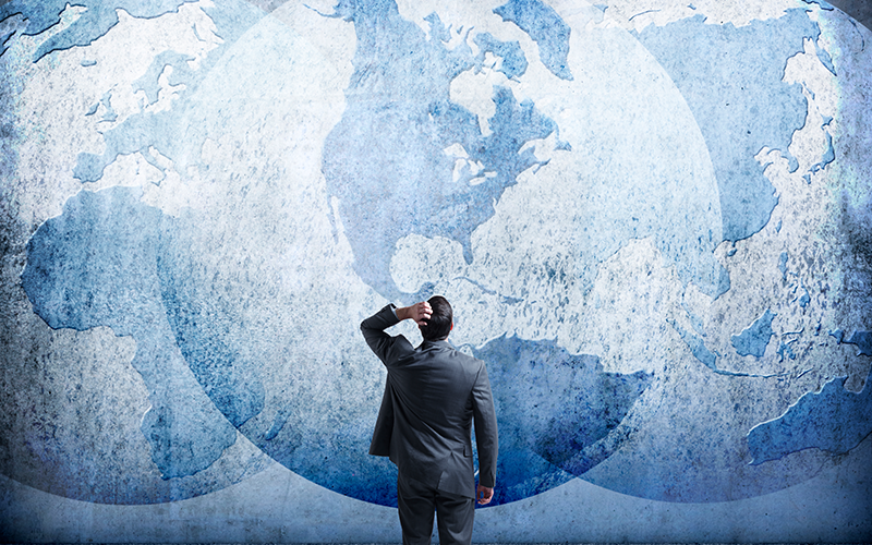 A rear view of a businessman as he stands and looks up at three interlocking globes on the wall in front of him as he tries to sort out the mess of geopolitical events.