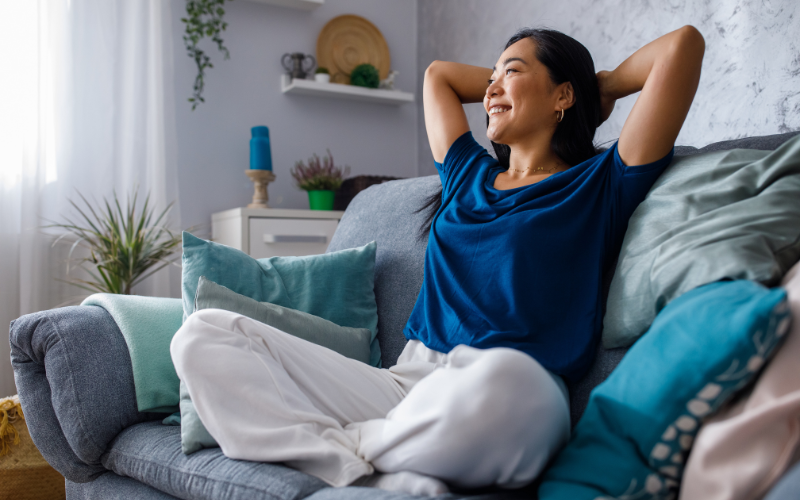 A woman sits on a sofa, smiling and leaning back with her hands behind her head