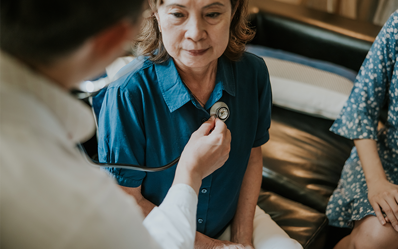 Asian Male doctor using a stethoscope to examining senior woman's lung and heartbeat during a healthcare home visit.