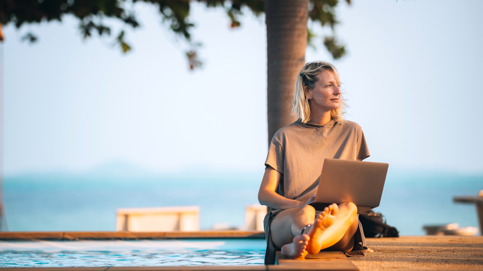 A woman sitting by the pool, working remotely with her laptop on her lap
