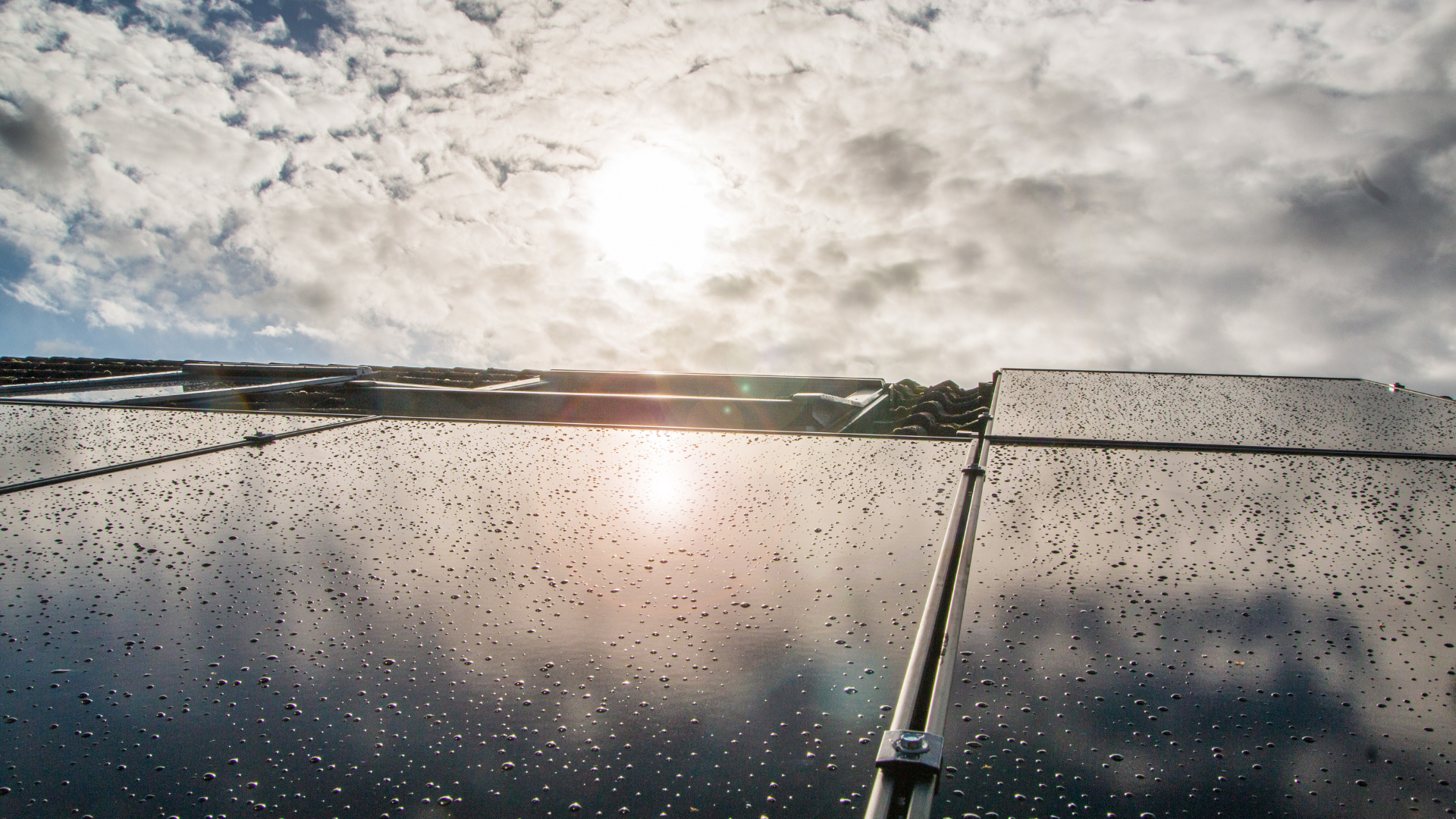 Solar panels glistening with raindrops under a partly cloudy sky, with the sun breaking through.