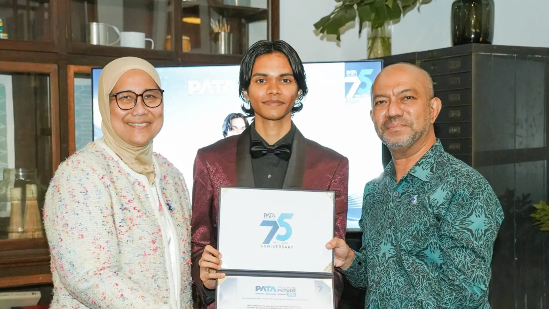 Abdulla Iyaan Ahmed holding his PATA Face of the Future 2026 Student Excellence certificate, flanked by two presenters in front of a PATA 75th anniversary screen