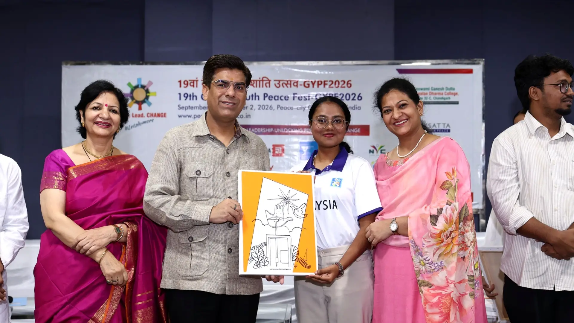 Mirsha Naresh Kumar stands with three other attendees, holding a framed illustration in front of the 19th Global Youth Peace Festival 2026 banner