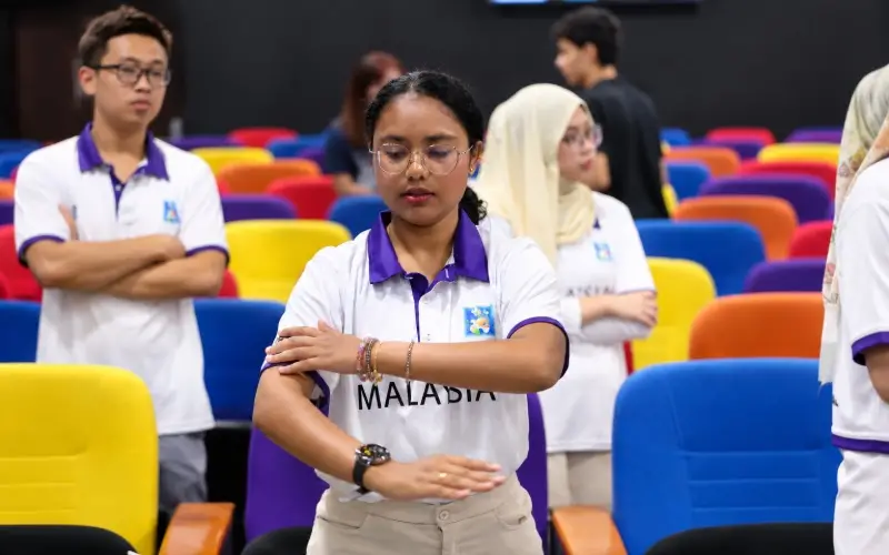 Mirsha Naresh Kumar demonstrates a hand movement while other participants stand among colourful auditorium seats