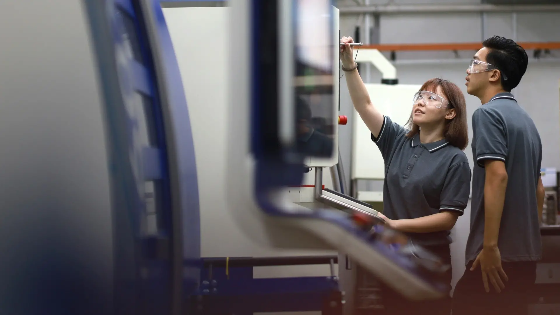 Two engineering students in safety glasses working at the control panel of a CNC machine