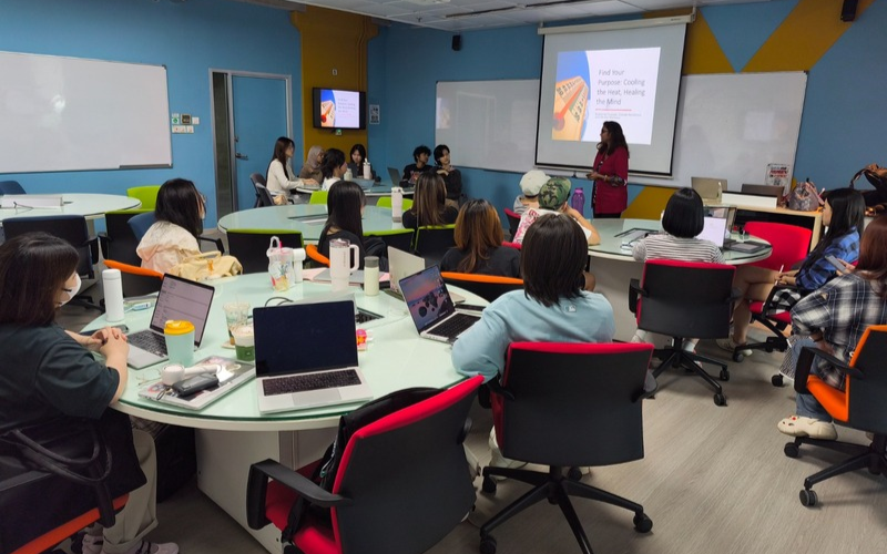 Dr Praveena stands at the front of a classroom giving a presentation while a group of students sit around round tables with laptops and notebooks, watching a projected slide on a screen.