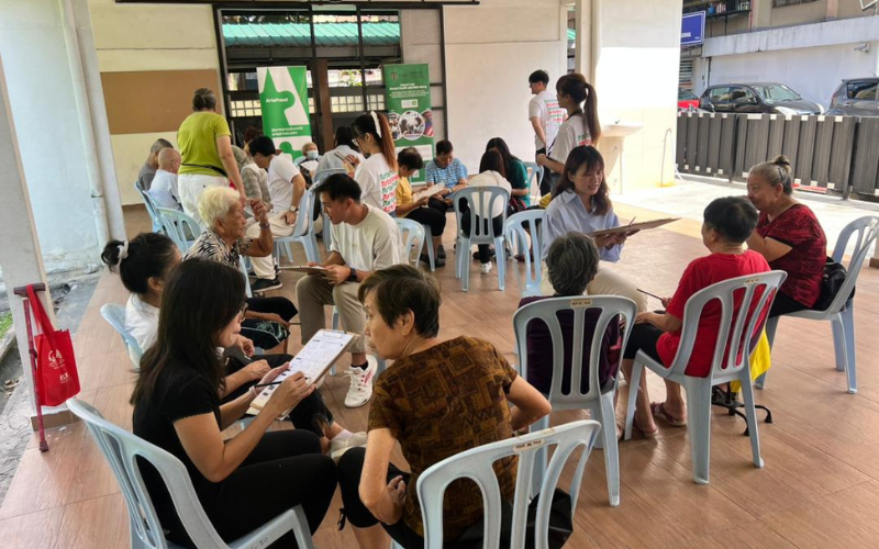 Several volunteers sit with elderly people in small groups on plastic chairs at an outdoor covered area, talking and filling out forms on clipboards.