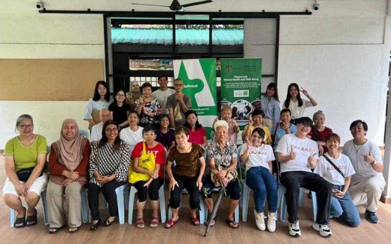 A large group of volunteers and elderly participants pose together for a group photo in a covered community space..