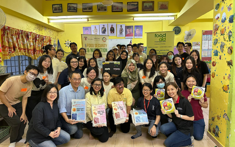 A large group of volunteers and organizers pose together inside a brightly painted yellow classroom during a Food Aid Foundation activity focused on food security and nutrition.