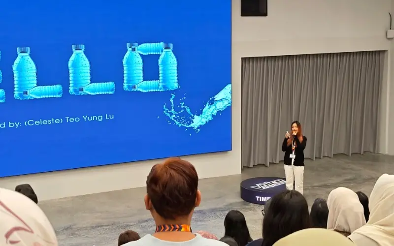 Celeste presenting on stage to a seated audience, beside a large screen showing her slide