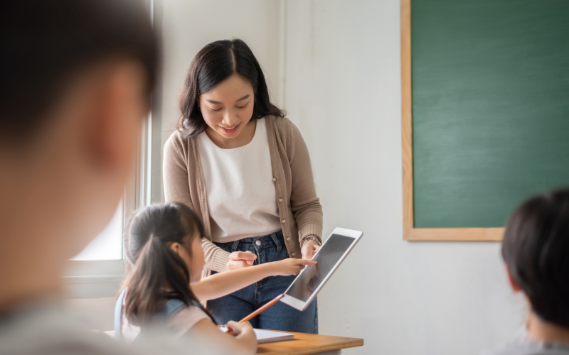 Asian female teacher teaching girl learning with digital tablet in classroom at primary school.
