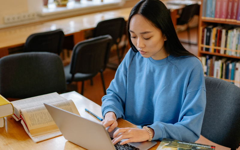 A student researching on a laptop at a library table, surrounded by open books and notes.