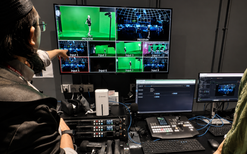 A crew member in the control room pointing at a multiview monitor showing the green-screen preview alongside the composited virtual set