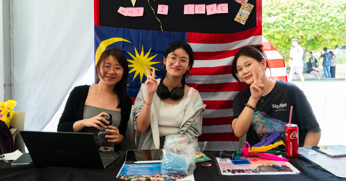 Three girls posing for the camera in front of a Malaysian flag