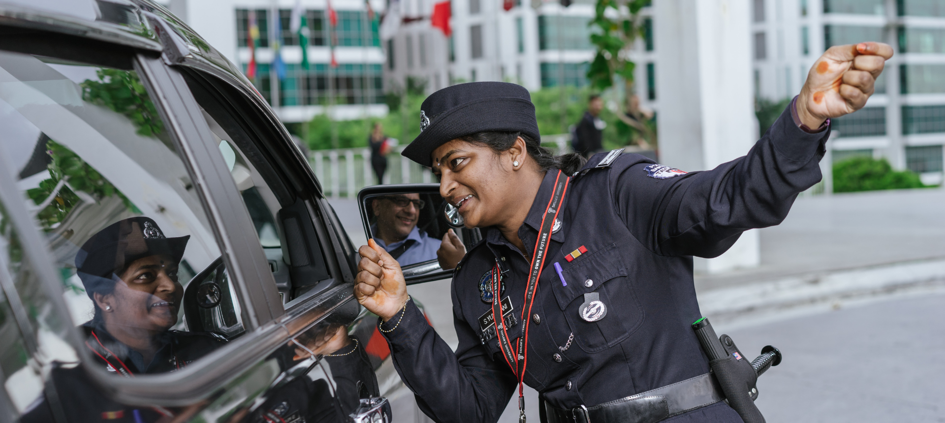 A smiling female security guard in uniform leans towards a car and gestures enthusiastically while interacting with the driver at a campus entrance.