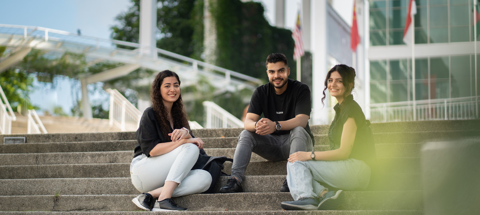 Three international students smile warmly as they sit together on wide, concrete steps on campus.