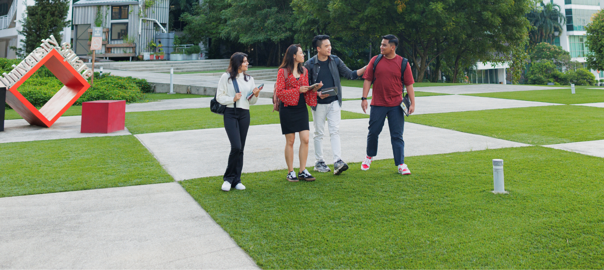 A group of four students socialising on a spacious lawn in front of large, modern campus buildings.