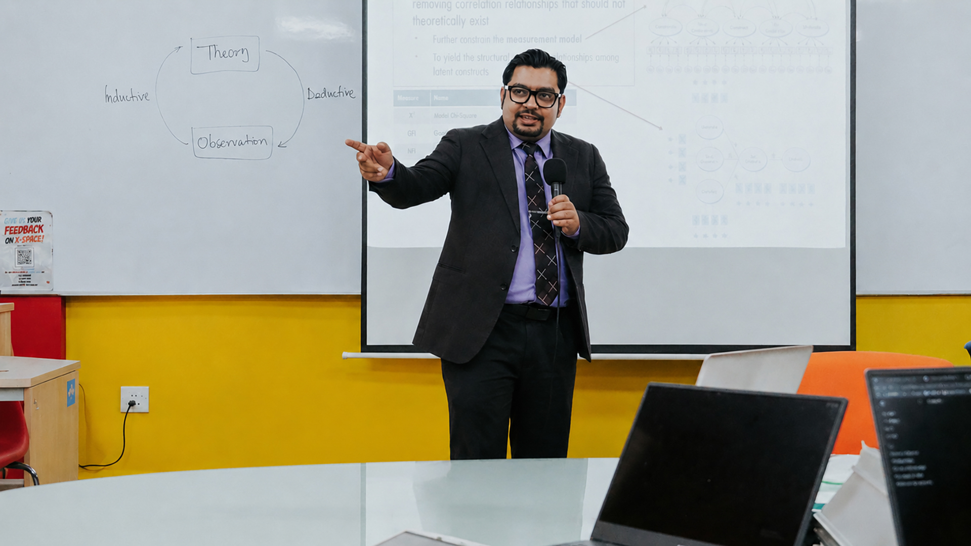 A lecturer in a suit holds a microphone and points toward a student while teaching, with a whiteboard and projected slides behind him and students seated with laptops in the foreground.