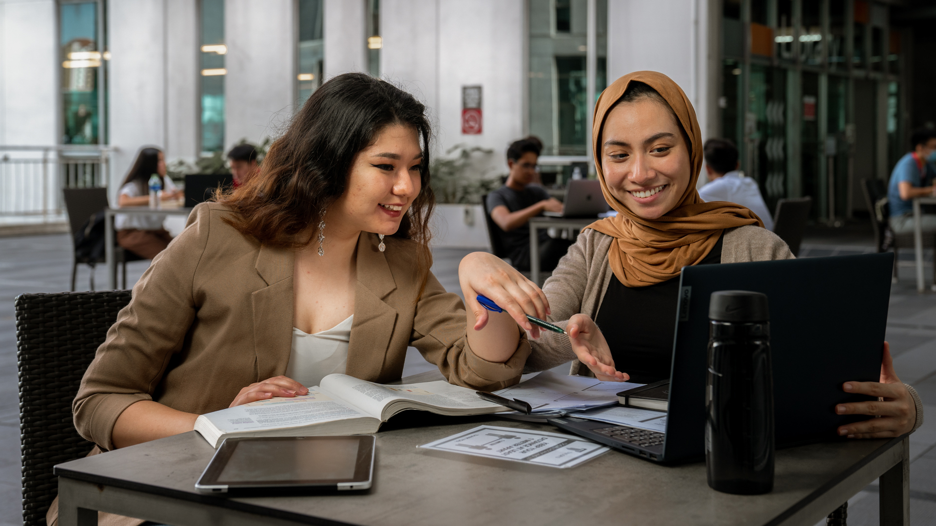 Taylor's students sit at an outdoor table studying together, smiling and discussing work while looking at a laptop, with books, a tablet, and notes spread out in front of them