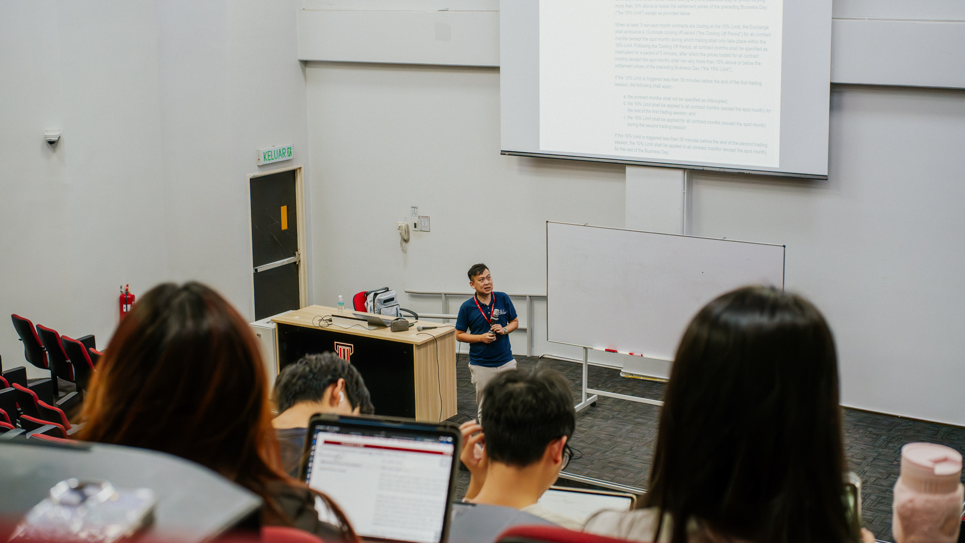 A lecturer stands at the front of a classroom speaking to students, with a whiteboard beside him and students in the foreground using laptops.