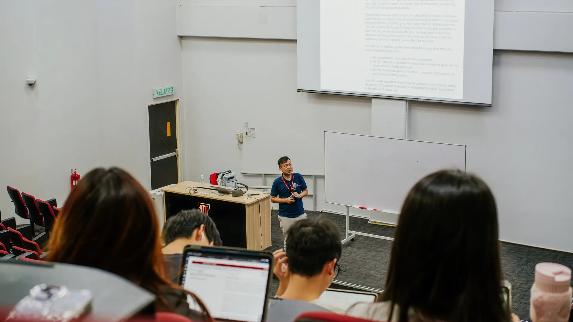 A lecturer stands at the front of a classroom speaking to students, with a whiteboard beside him and students in the foreground using laptops.