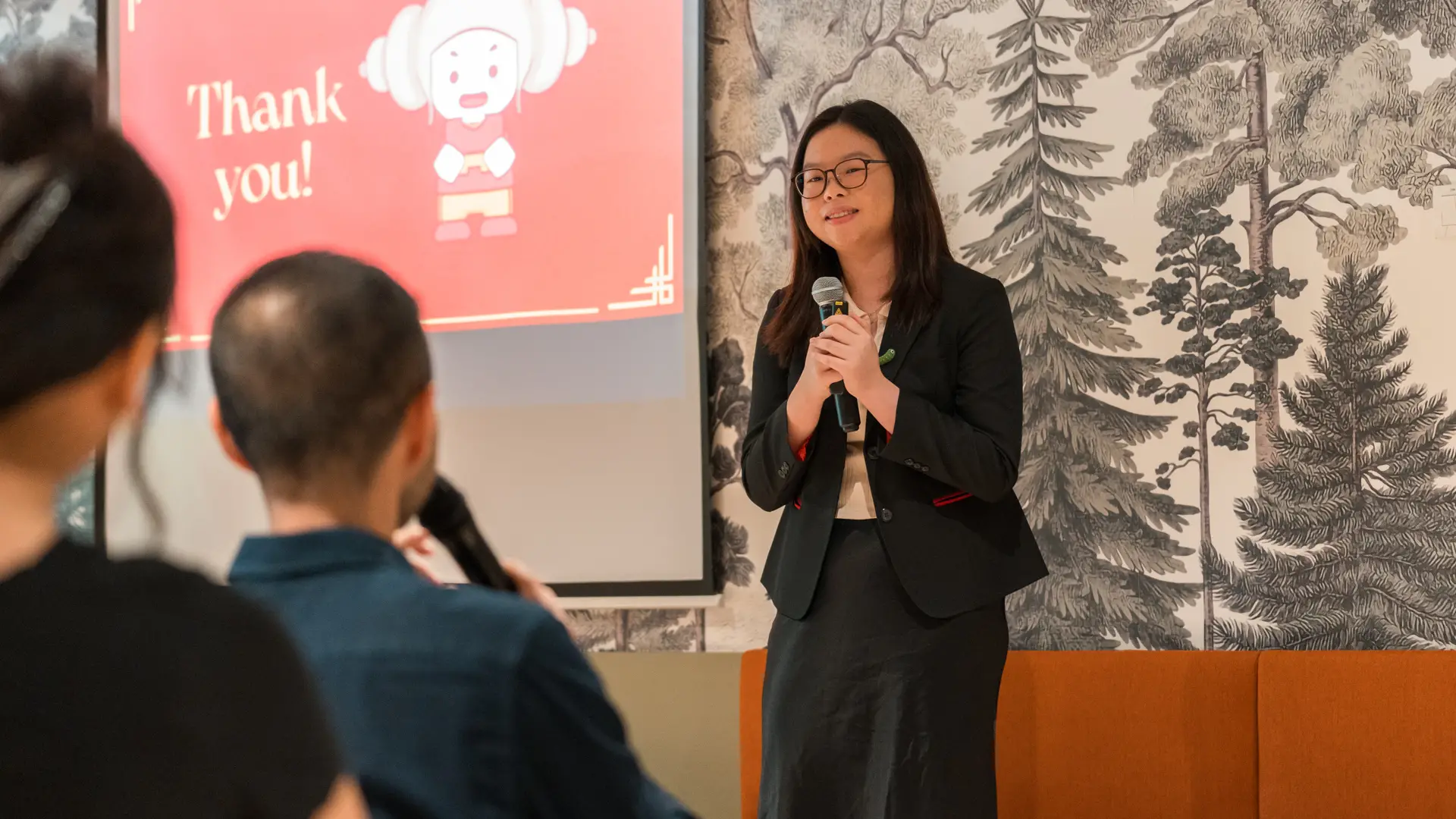 A student in business attire stands holding a microphone and speaking in front of an audience, with a presentation slide displayed behind her and a patterned wall as the backdrop