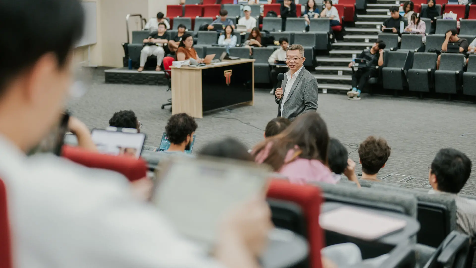 A lecturer holding a microphone speaks to students in a large lecture hall, with tiered seating filled with attendees and some students using laptops.