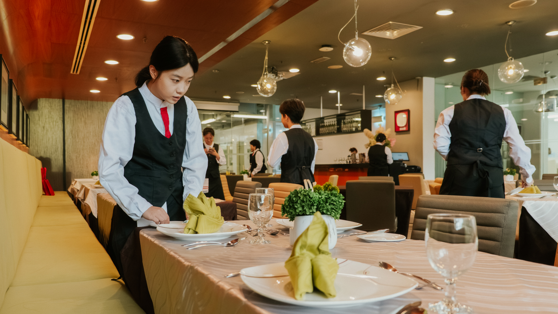 A Taylor's student carefully sets a dining table in a restaurant training environment, while other students prepare tables and service areas in the background under warm indoor lighting.